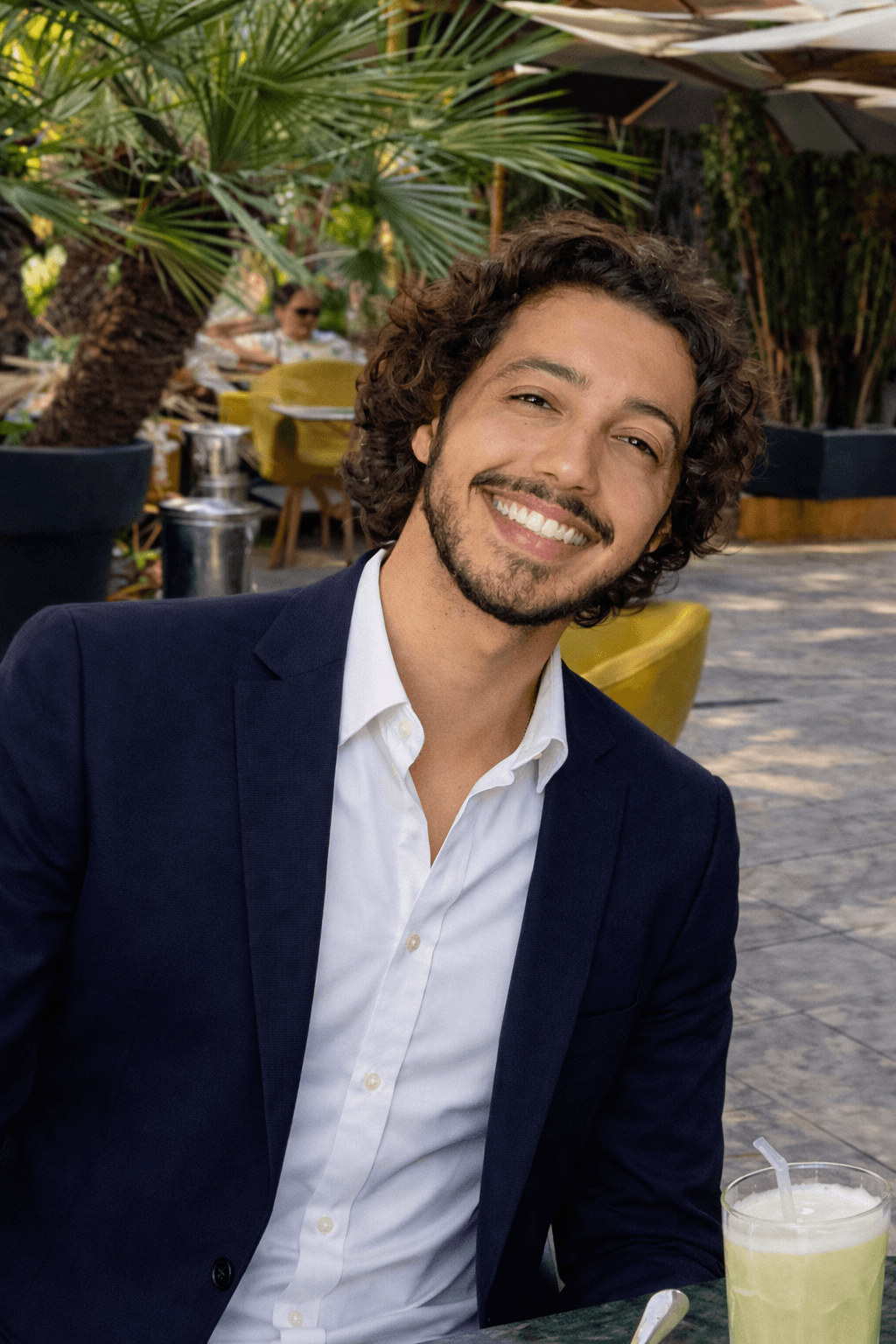 Homme souriant aux cheveux bouclés et barbe, portant un blazer bleu marine en terrasse.